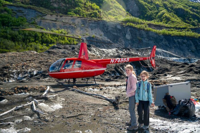 Two kids standing in front of a helicopter on a glacier, experiencing an exciting Alaskan adventure.