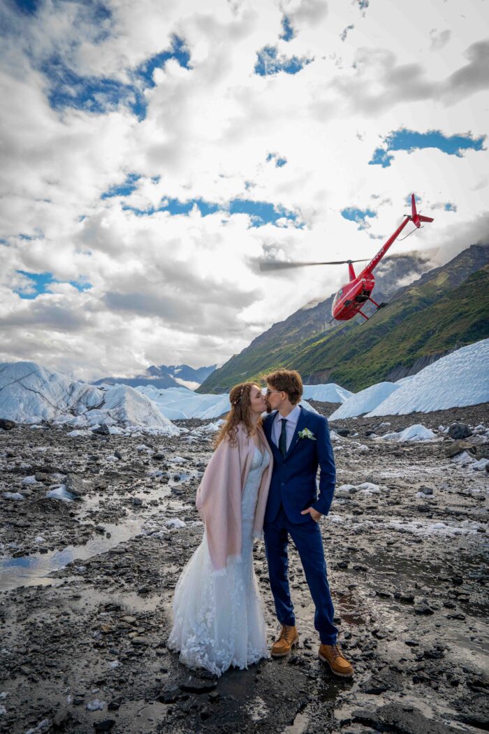 Bride and groom posing in front of a helicopter on a glacier, celebrating a unique Alaskan wedding adventure."