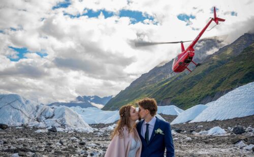 Bride and groom posing in front of a helicopter on a glacier, celebrating a unique Alaskan wedding adventure."