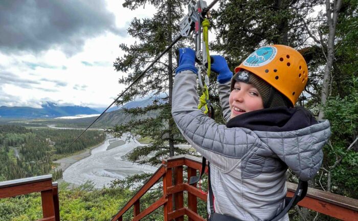 A kid smiling with excitement at the starting platform, ready to launch on an Alaskan zipline adventure.