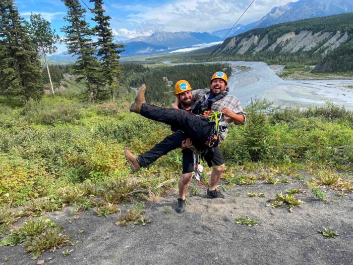 Two friends laughing and gearing up on the platform, ready for an epic zipline adventure in Alaska.